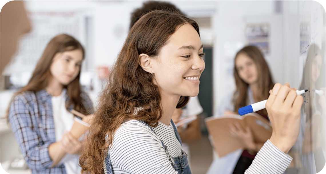 Young person writing on a whiteboard in the classroom while surrounded by other students. 