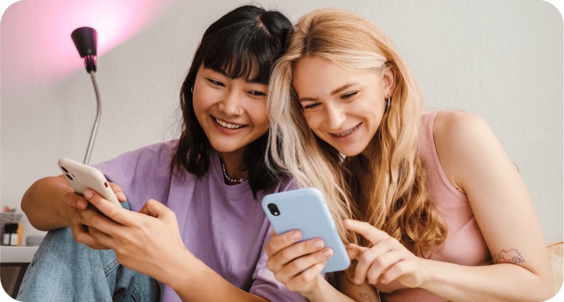 Two young people in a bedroom looking at their cell phones while smiling. 