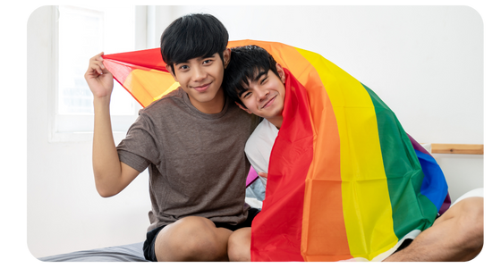 Two young Asian queer individuals cuddle holding a rainbow flag